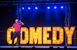 comedian standing on stage in front of large neon comedy sign