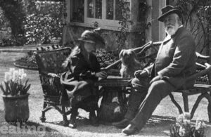 An old photograph of a young girl and an older man sitting on benches in a garden, with a dog nearby