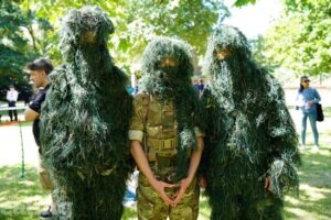 3 armed forces representatives standing in grass covered camo