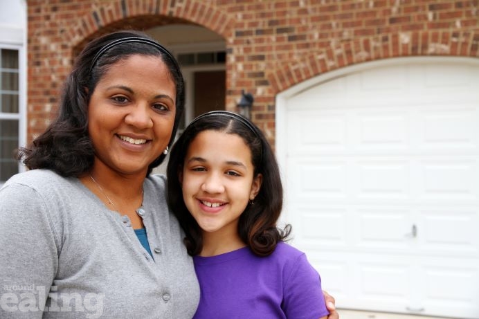A woman and a girl of about 11 years old standing together, smiling outside a house.