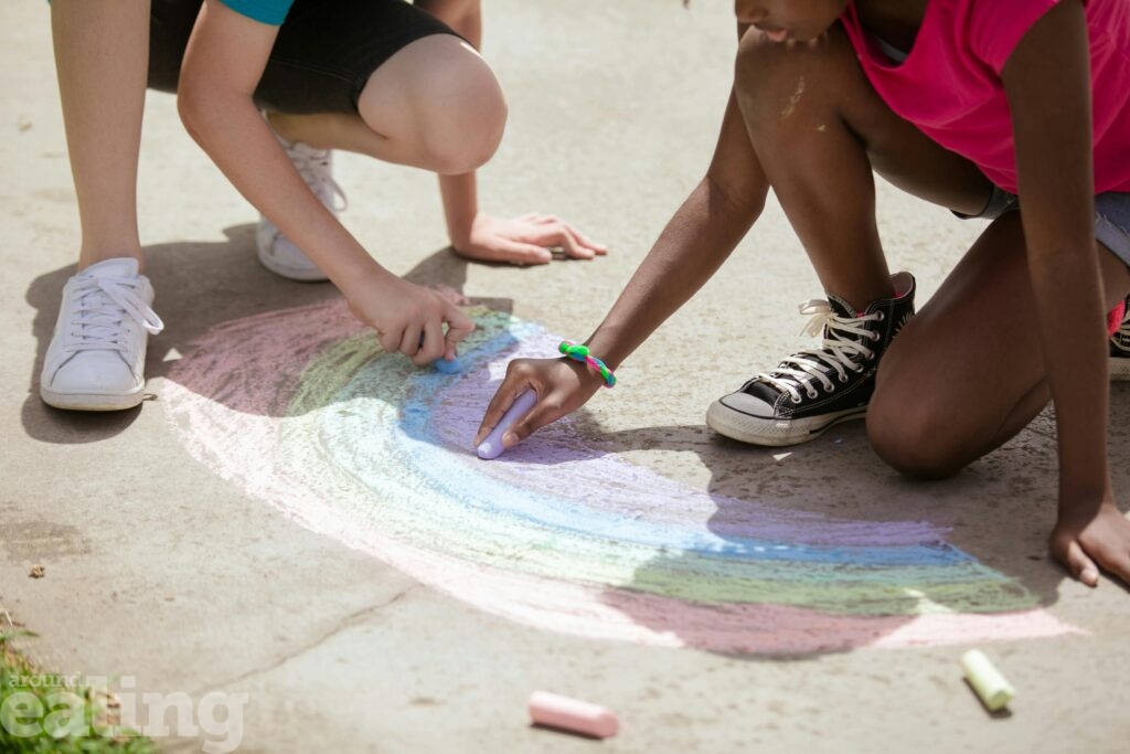 Two children drawing a rainbow on the street with chalk.