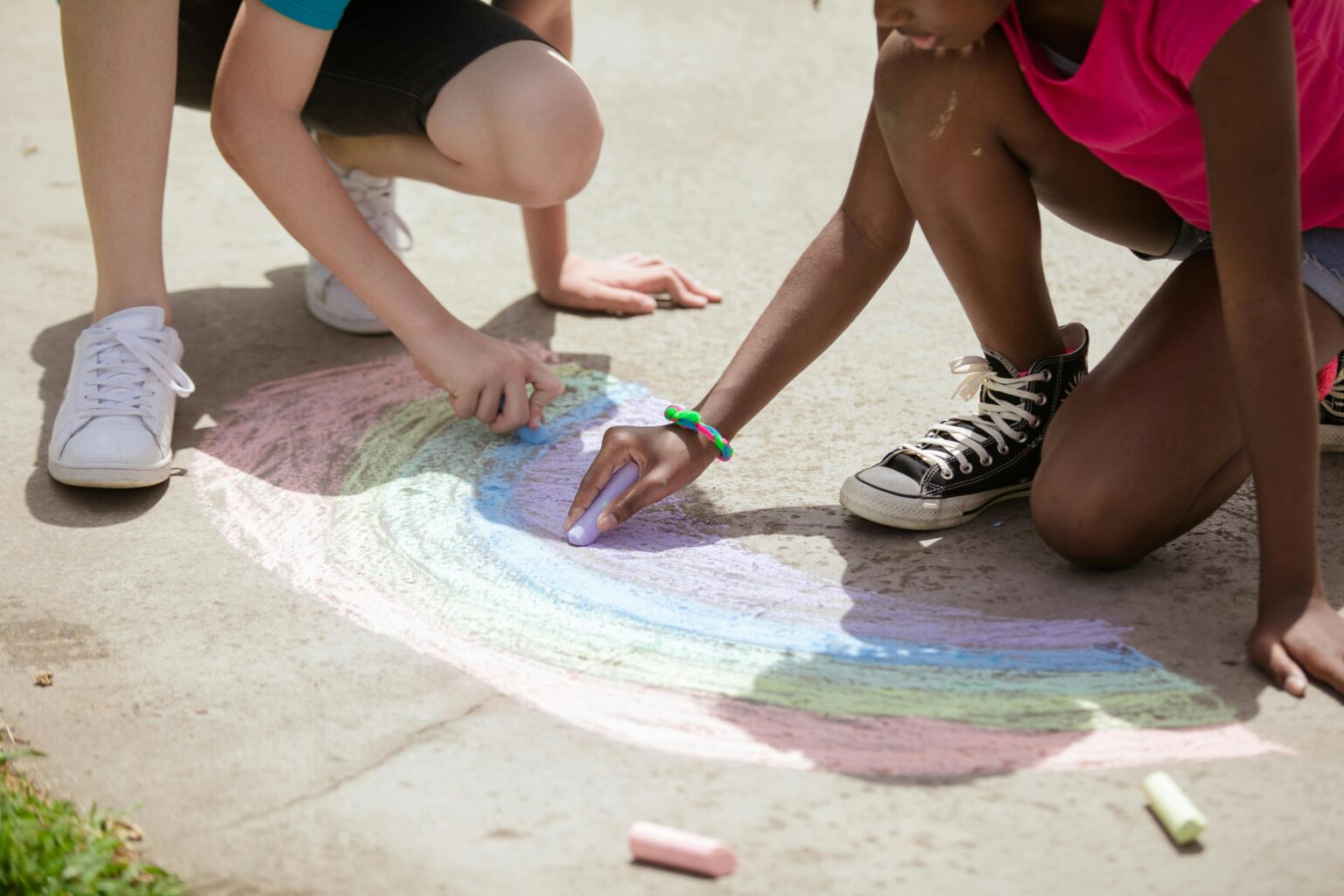 Two children drawing a rainbow on the street with chalk.