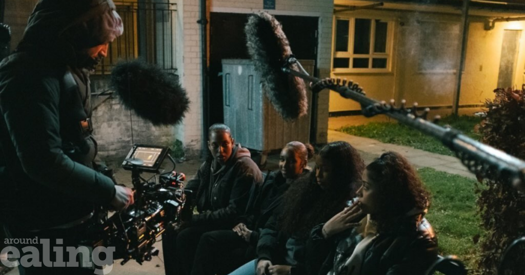 A group of young people sit on a bench being filmed