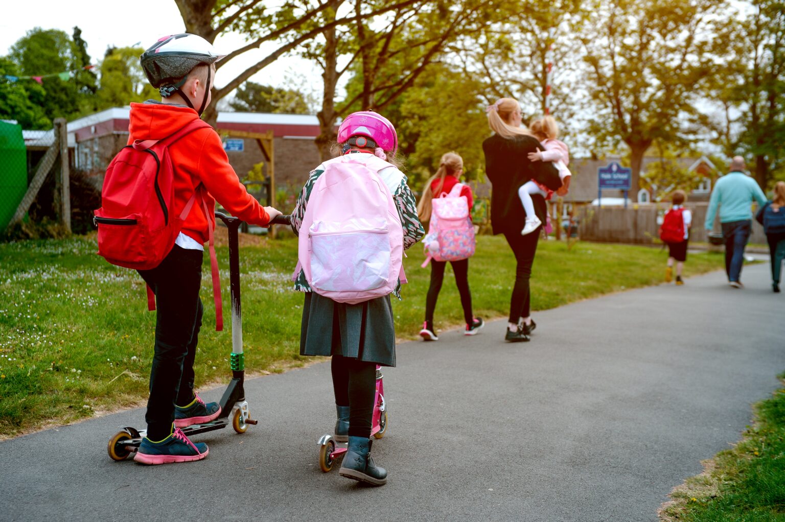 a woman carrying a child in front of children scooting on the pavement