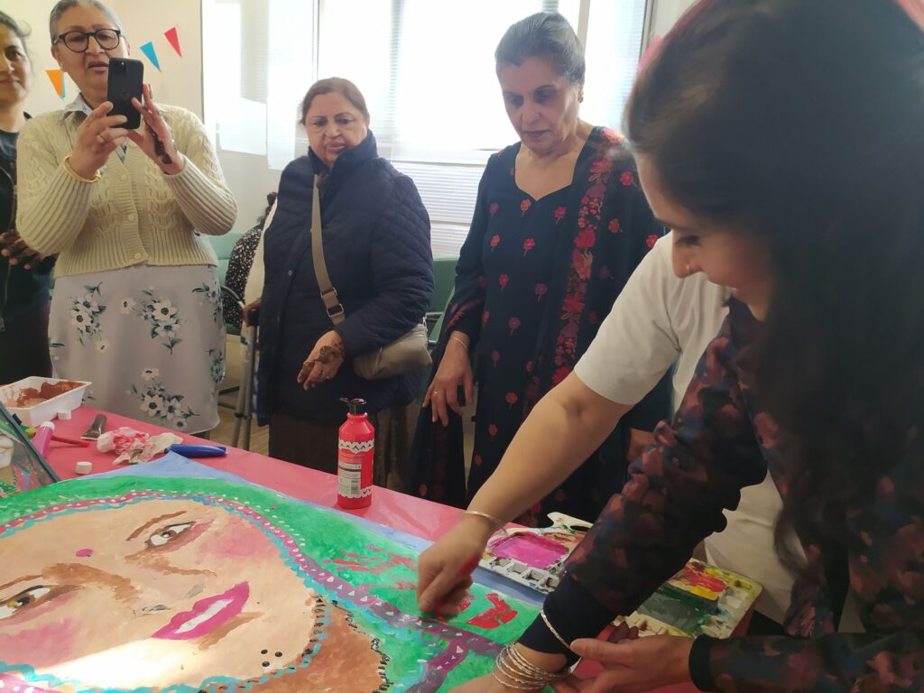 A group of women stand around table hand painting a large painting of a woman in a headscarf