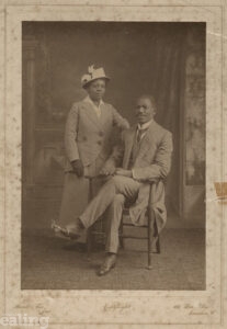 Man sitting on a chair, with woman standing by his side. Both dressed smartly. Black and white photo