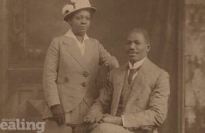 Man sitting on a chair, with woman standing by his side. Both dressed smartly. Black and white photo