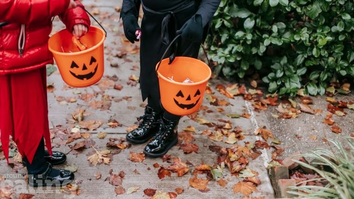 The legs and feet of two children dressed in Halloween costumes holding orange buckets with smiley faces.