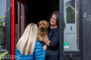 female resident at open door, holding her dog, talking to a woman on her doorstep holding phone and pen