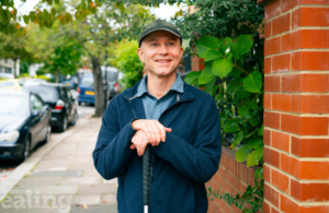 Man in cap stood smiling and facing the camera with both hands resting on the top of walking stick