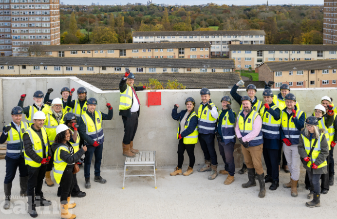 Group of people wearing high-vis jackets and construction helmets clapping and cheering, celebrating the topping out ceremony for new homes in Southall