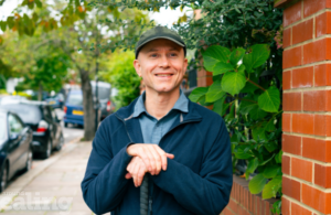 Man in cap stood smiling and facing the camera with both hands resting on the top of walking stick