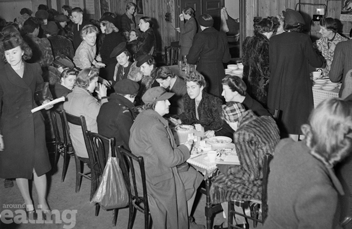 Table in a canteen in London in 1943, filled with people wearing coats and hats, eating food