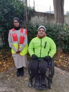 Huda stands next to Kudlip who sits in his wheelchair in a park.