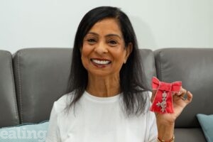 Shakuntla sitting on sofa ta home smiling and holding up her MBE medal