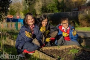 3 children kneeling at a flower bed outdoors, holding gardening equipment